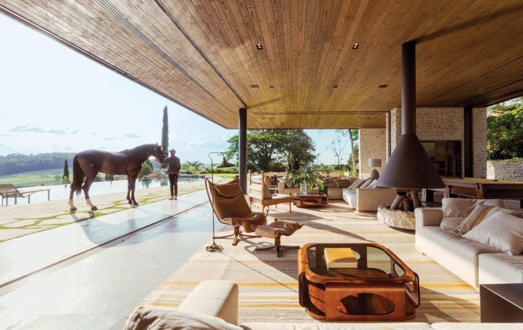 On either side of the sitting area’s freestanding fireplace, Casas-designed sofas anchor seating groups that include Sigurd Ressel’s Falcon lounge chair and ottoman on the left, Sergio Rodrigues’s Tonico armchair behind it, and a pair of jacaranda coffee tables by Jorge Zalszupin; to the right, there’s a games area with a pool table.